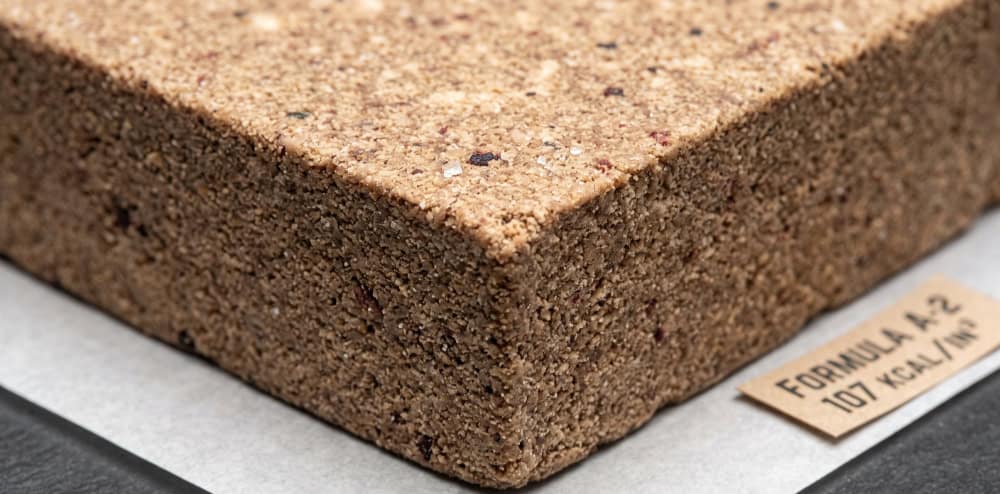 Close-up of a dense, compressed survival ration block with a coarse, tightly packed texture, beside a card reading Formula A-2, 107 kcal per cubic inch.